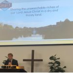 Pastor Chris Riser sharing podium with powerpoint slide showing Grace Community Church logo and caption Sharing the unsearchable riches of our Lord Jesus Christ in a dry and thirsty land.