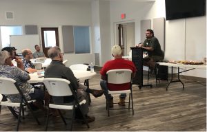 Jim Quigley teaching at podium to a small group of men seated in folding chairs at round tables